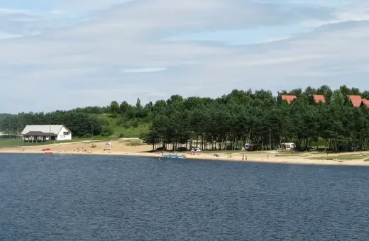 Słoneczny dzień nad jeziorem Chańcza. Piękna plaża, las i kilka domków w tle. Ludzie odpoczywają i kąpią się.