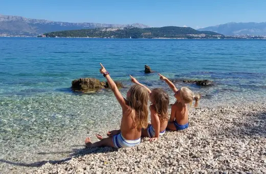 Trzy dziewczynki siedzą na kamienistej plaży w Chorwacji, wskazując palcami na morze. Idealne miejsce, gdzie do Chorwacji z dziećmi.