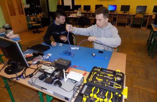 Dwóch uczniów ćwiczy technikę informatyczną, montując kable sieciowe. Na stole sprzęt i narzędzia.