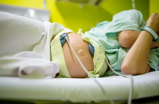 Kobieta w ciąży leży na łóżku, podłączona do aparatury KTG. Widać jej brzuch z przyczepionymi czujnikami.