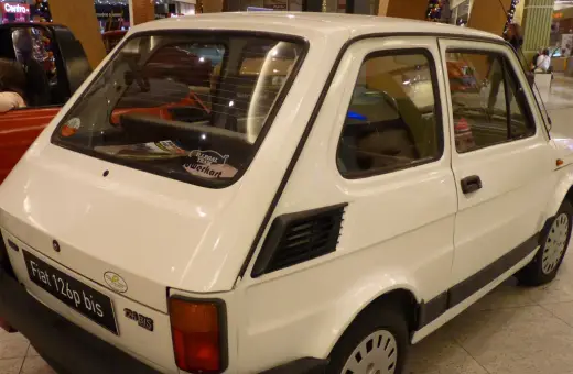 Biały Fiat 126p bis z naklejkami na tylnej szybie, stojący obok czerwonego samochodu.