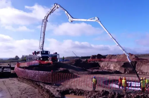 Ogromna pompa do betonu pracuje na budowie, dostarczając materiał do wykopu. Pracownicy w kamizelkach obserwują proces.