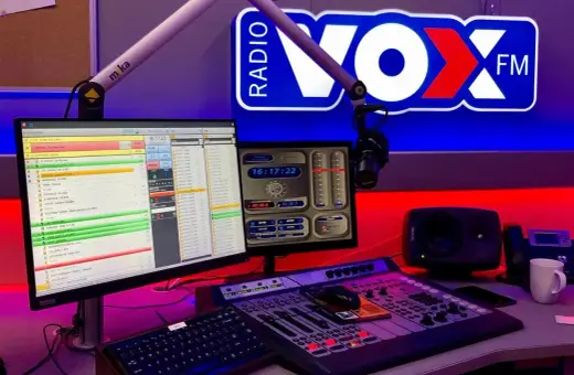 Studio radiowe VOX FM Warszawa. Komputery z listą utworów, mikser, mikrofon i logo stacji.
