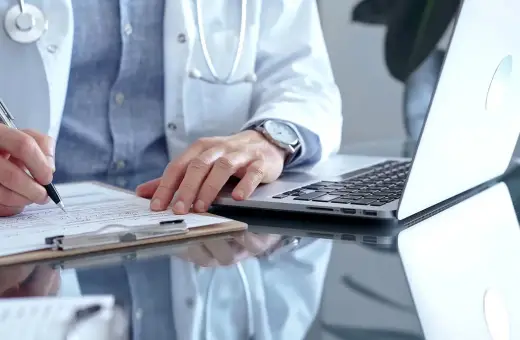 Lekarz w białym kitlu pisze na dokumentach, obok laptop. Szybka konsultacja psychiatryczna online.