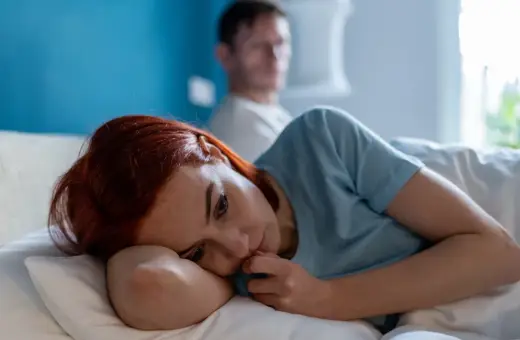 Smutna kobieta leży w łóżku, zamyślona. W tle niewyraźny mężczyzna. Obraz symbolizuje trudne relacje i toksyczny partner cechy.