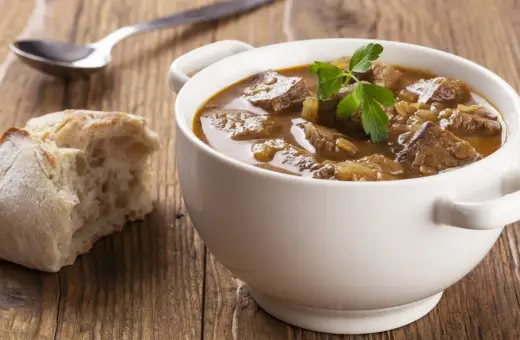 Aromaticzny, tradycyjny gulasz wołowy w białej miseczce, z gałązką pietruszki, obok kromka chleba i łyżka.