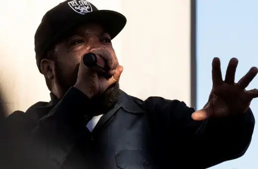 Raper Ice Cube w czapce z daszkiem "ICE CUBE" śpiewa do mikrofonu, gestykulując ręką. Jego styl to klasyczny diss.