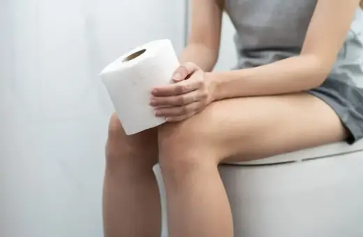 Kobieta siedzi na toalecie, trzymając rolkę papieru toaletowego. Zastanawia się, czy niepokojący zapach amoniaku w moczu to objaw problemu.