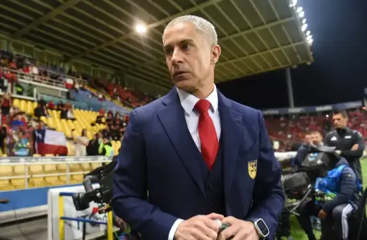 Sylvinho, trener w granatowym garniturze i czerwonym krawacie, stoi na tle trybun stadionu z flagą Chile.