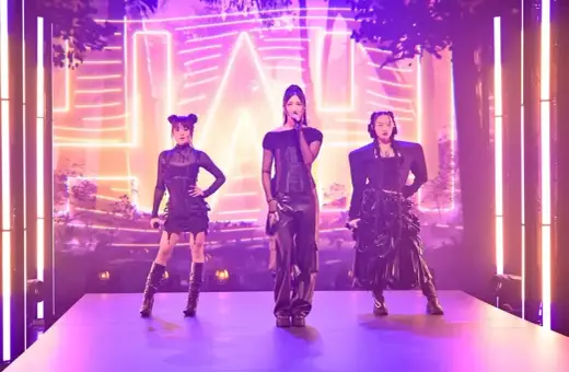 Trzy artystki k-pop w czarnych, stylizowanych strojach na scenie z neonowym tłem.