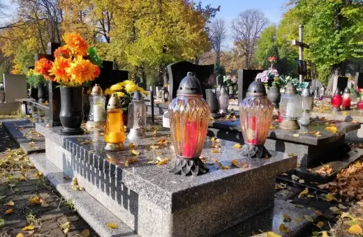Jesienny dzień na cmentarzu. Na kamiennym, murowanym grobie palą się znicze, a obok stoją świeże kwiaty.