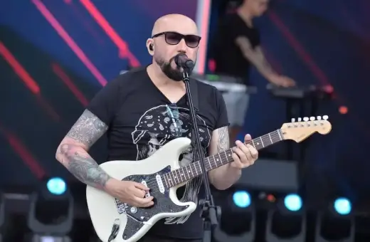 Artysta z tatuażami gra na białej gitarze, śpiewając do mikrofonu. W tle widać klawiszowca. To z pewnością niezapomniane **kombi utwory** na żywo!