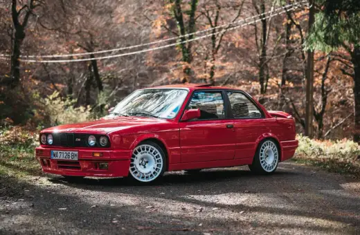 Czerwone bmw e30 coupe z białymi felgami na tle jesiennego lasu.
