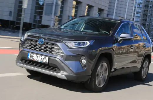 Ciemnoszary Toyota RAV4 Hybrid pędzi po mieście, odbijając w lustrach budynki i niebo.