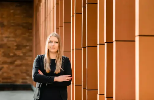 Kobieta w czarnym garniturze stoi na tle ceglanej ściany i pomarańczowych pionowych elementów.