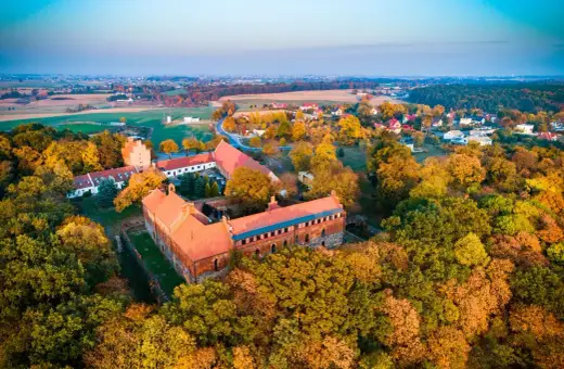 Zamek Bierzgłowski otoczony jesiennym lasem, z czerwonymi dachami i ceglanymi murami, w blasku zachodzącego słońca.
