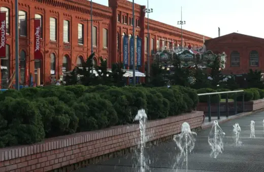 Piotrkowska Łódź: ceglane budynki Manufaktury, karuzela, fontanny i zielone krzewy tworzą letni klimat.