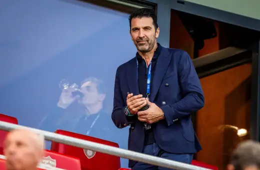 Gianluigi Buffon, legendarny włoski bramkarz, w eleganckim granatowym garniturze, obserwuje mecz z trybun.