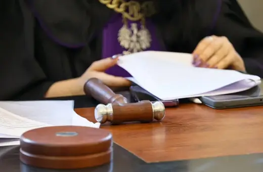 Sędzia w todze z orłem przegląda dokumenty. Na stole młotek i telefony. Sprawa spadkobiercy w toku.