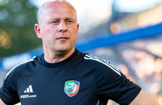 Łysy mężczyzna w czarnej koszulce z herbem Miedzi Legnica. Trener Miedzi Legnica w skupieniu obserwuje boisko.