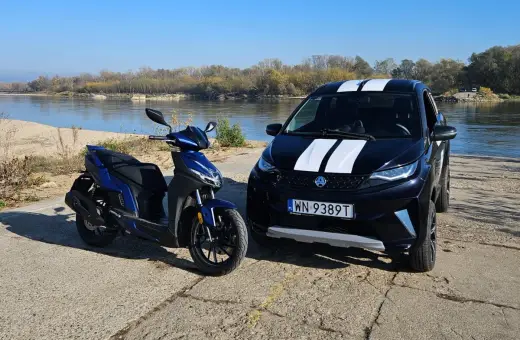 Niebieski skuter i czarne aixam auto z białymi pasami stoją nad rzeką.