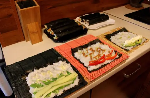 Przygotowanie ryżu do sushi. Na matach leżą arkusze nori z ryżem i dodatkami: ogórkiem, awokado, papryką i sosem.