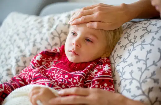 Małe dziecko w czerwonym sweterku z wzorem płatków śniegu leży chore, z gorączką. Ręka opiekuna sprawdza temperaturę. To typowy obrazek, gdy dopada nas wirus.