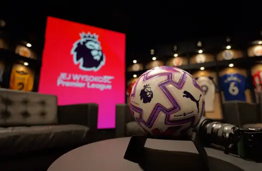 Piłka do gry w Premier League na tle ekranu z logo ligi. W tle widoczne koszulki piłkarskie.