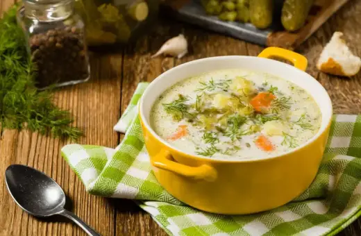 Zupa ogórkowa z koperkiem, ziemniakami i marchewką. W tle słoik z pieprzem, czosnek i ogórki kiszone. Idealne jakie przyprawy do zupy ogórkowej?