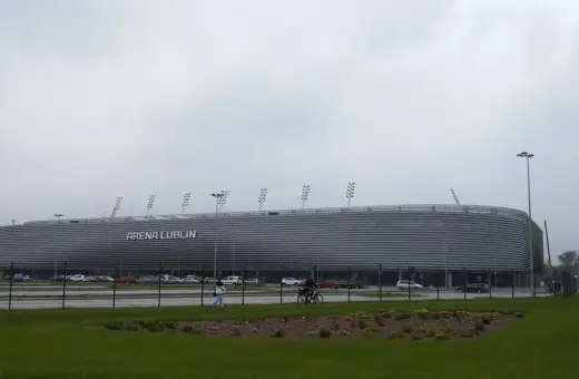 Nowoczesna Arena Lublin z napisem "ARENA LUBLIN" na fasadzie. Obok parking, a na pierwszym planie trawnik. Pojemność obiektu robi wrażenie.