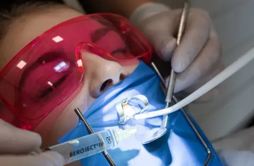 Precyzyjne leczenie kanałowe pod mikroskopem. Dentysta w rękawiczkach używa narzędzi i strzykawki. Pacjent ma założone różowe okulary ochronne.