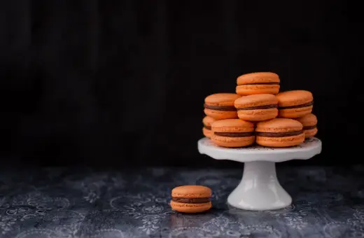 Pomarańczowe makaroniki francuskie z czekoladowym nadzieniem ułożone na białej paterze, jeden makaronik leży obok.