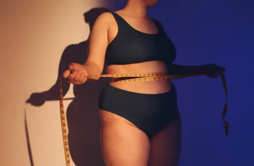 Osoba mierzy talię centymetrem. Ciemne światło podkreśla kształty, wywołując dyskomfort związany z body shaming.