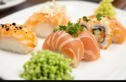 Talerz z różnorodnym nigiri sushi z łososiem, posypane sezamem i ozdobione natką pietruszki. Obok porcja zielonego wasabi.