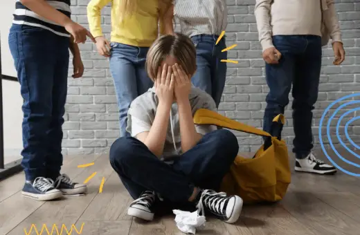 Chłopiec siedzi na podłodze, zakrywając twarz dłońmi, podczas gdy grupa dzieci stoi nad nim. To obraz tego, czym jest **bullying**.