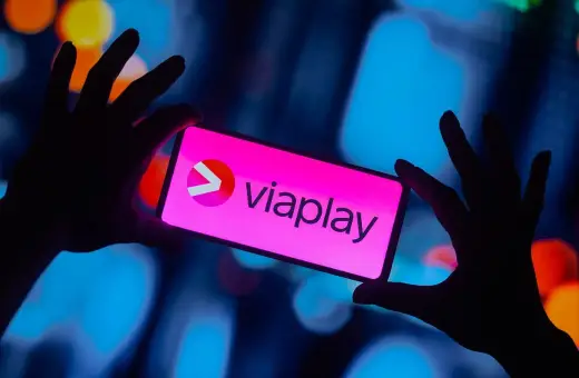 Dłonie trzymają smartfon z logo Viaplay. W tle rozmyte, kolorowe światła.
