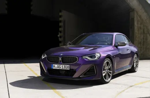 Fioletowe BMW 220i coupe z nowoczesnym designem, zaparkowane na betonowym placu przed ciemnym budynkiem.