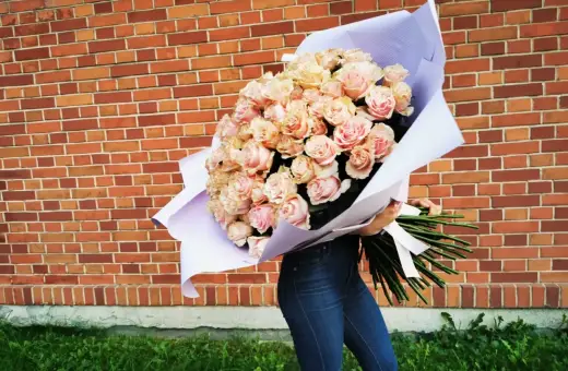 Ogromny bukiet 108 róż w odcieniach różu i brzoskwini, owinięty w fioletowy papier, trzymany przez osobę w dżinsach na tle ceglanej ściany.