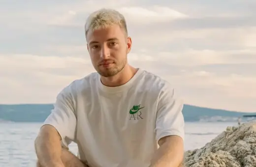 Edzio rap, z blond włosami i w białej koszulce, siedzi na plaży.