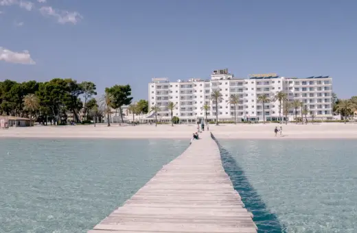 Drewniany pomost prowadzi do piaszczystej plaży Playa de Muro z białym hotelem i palmami w tle.