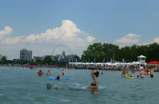 Słoneczna balaton plaża tętni życiem. Ludzie kąpią się, pływają na materacach i odpoczywają pod parasolami. W tle widać diabelski młyn.