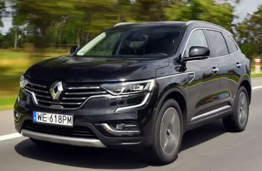Czarny Renault Koleos 2.0 dCi pędzi po drodze, otoczony rozmytym zielonym krajobrazem.