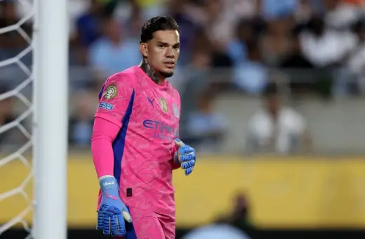 Bramkarz Manchester City w różowym stroju, z niebieskimi rękawicami, gotowy do obrony bramki.