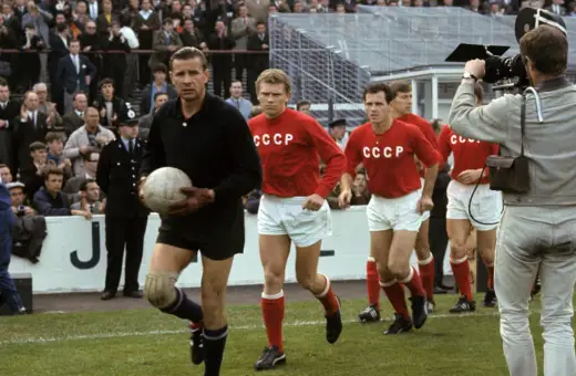 Bramkarz w czarnym stroju trzyma piłkę, obok niego biegną zawodnicy w czerwonych koszulkach z napisem CCCP. Kamera filmowa rejestruje mecz.