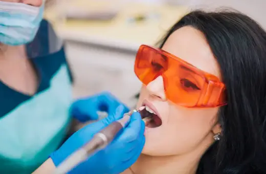 Pacjentka w okularach ochronnych poddaje się zabiegowi stomatologicznemu. Dentysta w rękawiczkach używa wiertła. Nowoczesne znieczulenie komputerowe zapewnia komfort.
