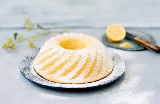 En vacker citronkaka i långpanna, pudrad med florsocker, står på ett fat. En halv citron och en kniv syns i bakgrunden.