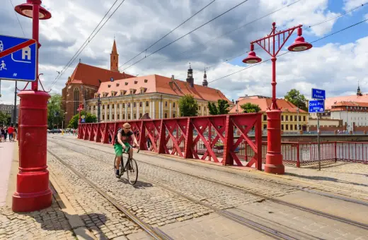 Mężczyzna na rowerze jedzie po brukowanej drodze rowerowej z torami tramwajowymi, w tle zabytkowe budynki i czerwony most.