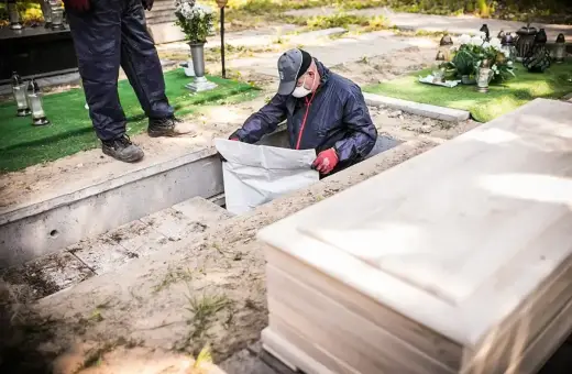 Ekshumacja zwłok po 40 latach. Pracownicy w masce i rękawiczkach wyciągają worek z grobu. Obok leży nowa trumna.