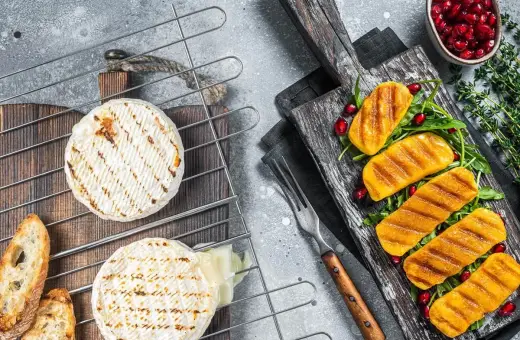 Dwa okrągłe sery camembert grillowane na ruszcie, obok pieczywo i grillowane kawałki sera na desce z granatem.