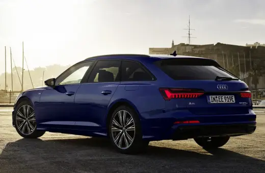 Niebieskie Audi A6 Avant z napisem "50 TFSI e" na klapie bagażnika, zaparkowane przy nabrzeżu z widokiem na zabytkową fortecę i żaglówki.